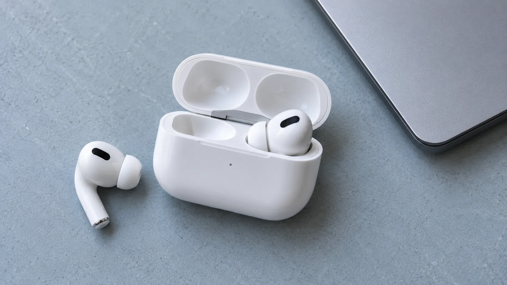 Close product photograph of white wireless earbuds and their open case beside a silver laptop edge.